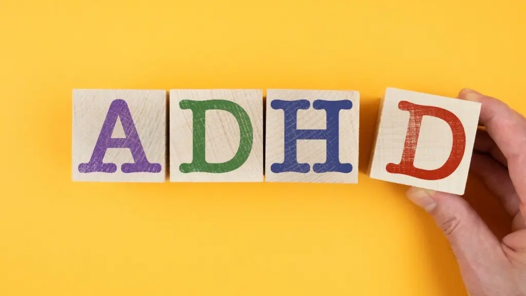 Adult ADHD nutrition concept featuring colorful wooden blocks spelling “ADHD” against a bright yellow background, with a hand holding the final letter.
