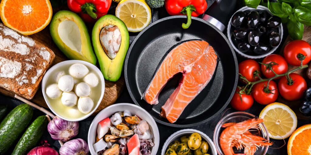 Fresh foods commonly featured in the MIND Diet vs Mediterranean Diet, including salmon, avocado, olives, vegetables, citrus, seafood, and whole grain bread. The MIND, Mediterranean, and DASH diets emphasize these nutrient-rich foods for brain and heart health.