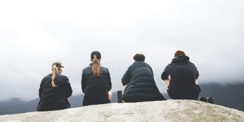 Four people sitting together on a rocky hillside overlooking misty mountains, illustrating social connection and mental wellbeing in a discussion about the causes of mental health problems.